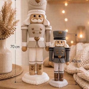 Könnte beinhalten: Zwei Nussknacker-Figuren auf einer Holzoberfläche. Der größere Nussknacker ist 70 cm groß, beige und weiß mit goldenen Herzakzenten. Der kleinere Nussknacker ist 40 cm groß, grau und weiß mit goldenen Herzakzenten verziert.