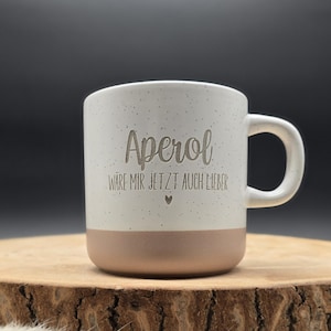 Könnte beinhalten: Ein weißer Keramikbecher mit gesprenkelter Oberfläche und einem glatten, beigefarbenen Boden. Auf der Vorderseite befindet sich der eingravierte Text "Aperol" über dem Satz "WÄRE MIR JETZT AUCH LIEBER" und einem kleinen Herzsymbol.