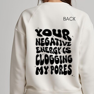 May include: A white sweatshirt with the text "YOUR NEGATIVE ENERGY IS CLOGGING MY PORES" printed on the back in black, bubble letters.