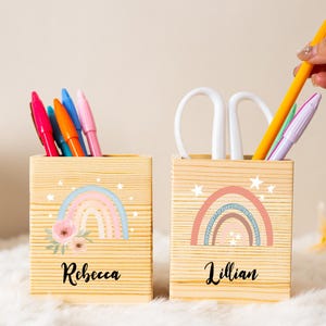 Op de afbeelding: Twee houten pennenbakjes met regenboog- en bloemmotieven. Eén is gemarkeerd met "Rebecca" en de andere met "Lillian". De houders bevatten pennen, een schaar en een potlood. De achtergrond is een zacht, wit, pluizig oppervlak.