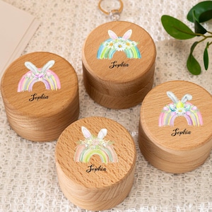 Puede incluir: Cuatro pequeñas cajas redondas de madera clara. Cada tapa presenta un diseño de arcoíris pastel con orejas de conejo y el nombre "Sophia". Una caja tiene un llavero.