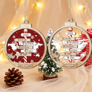 Pode incluir: Duas decorações de Natal em madeira com um design de mapa-múndi. Cada enfeite apresenta um esquema de cores vermelho e madeira natural, com o ano "2025" e placas de destino para Nova York, São Francisco, Nova Zelândia, Austrália e Sydney. Um também inclui Veneza. Os nomes "James & Amelila" estão inscritos.
