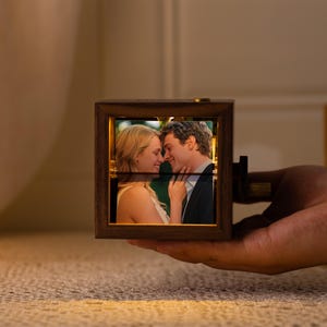 May include: A wooden photo cube with a picture of a couple. The cube is held in a hand and has a warm, glowing light inside. The frame is a dark brown color.