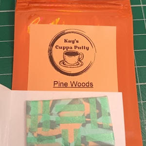 May include: An orange packet with a label displaying "Kay's Cuppa Putty" and "Pine Woods." Beneath the label is a small, rectangular item with a green, orange, and grey design. The background is a green cutting mat.