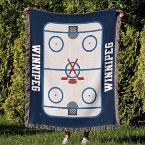 Puede incluir: Una manta azul marino con un diseño de pista de hockey. La palabra "WINNIPEG" está escrita en blanco a ambos lados. El centro es crema con círculos azules y palos de hockey rojos. Los bordes están deshilachados.