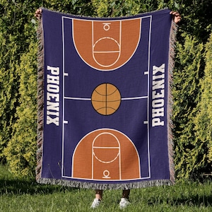 May include: A blue woven throw blanket with a basketball court design and the word "PHOENIX" on each side. The court is orange and brown with a basketball in the center.