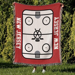 May include: Red woven throw blanket with a white hockey rink design and the words "NEW JERSEY" on either side.