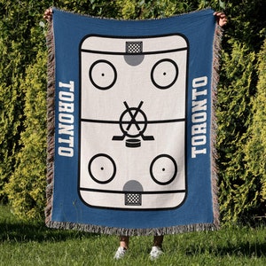 May include: A blue woven throw blanket with a white hockey rink design. The rink has two goals, a puck, and two hockey sticks crossed in the center. The words "TORONTO" are printed on both sides of the rink.