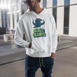 May include: White hooded sweatshirt with a graphic of a blue eagle wearing sunglasses and the words "WORLD CHAMPS" in green and blue. The sweatshirt has a front pocket and is being worn with blue jeans.