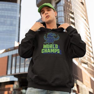 May include: Black hooded sweatshirt with a green and blue graphic of a bird and the words "WORLD CHAMPS". The sweatshirt has a front pocket and a drawstring hood. The person in the image is wearing a green cap and blue jeans.