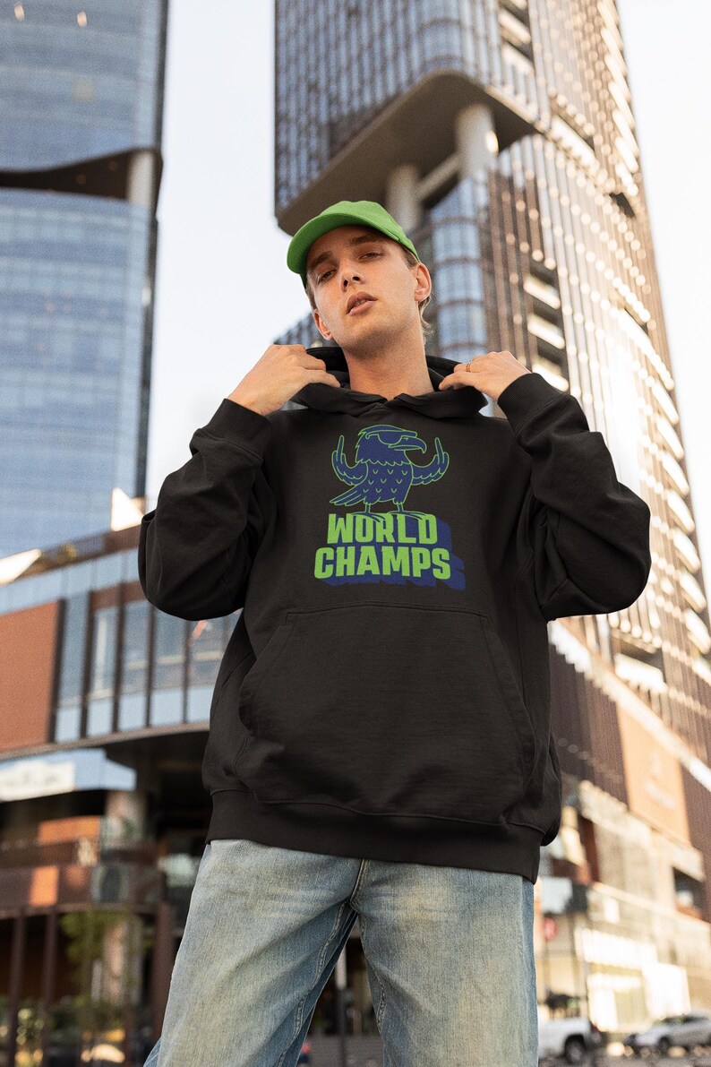 May include: Black hooded sweatshirt with a green and blue graphic of a bird and the words "WORLD CHAMPS". The sweatshirt has a front pocket and a drawstring hood. The person in the image is wearing a green cap and blue jeans.
