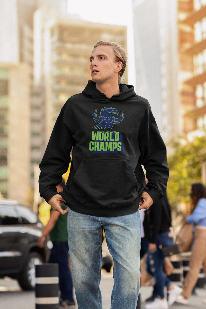 May include: Black hooded sweatshirt with a blue and green graphic of a bird wearing sunglasses and the words "WORLD CHAMPS". The sweatshirt has a front pocket and is worn with blue jeans. The image is taken outdoors.