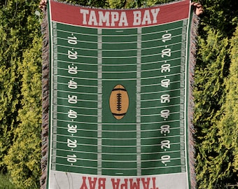 May include: A green and red woven throw blanket with the words "Tampa Bay" and a football field design. The blanket has fringe on all sides.