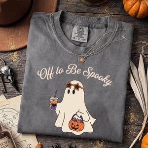 May include: A charcoal gray short-sleeve t-shirt featuring a graphic of a white ghost wearing star-shaped sunglasses, holding a colorful drink and a jack-o'-lantern bucket. The text "Off to Be Spooky" is printed in a cream-colored script font.