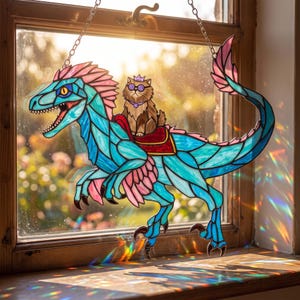 May include: A stained glass suncatcher featuring a blue and pink dinosaur with a brown cat wearing a purple crown and round glasses riding on its back. The piece is suspended by a metal chain and displays intricate glass segments and colours.