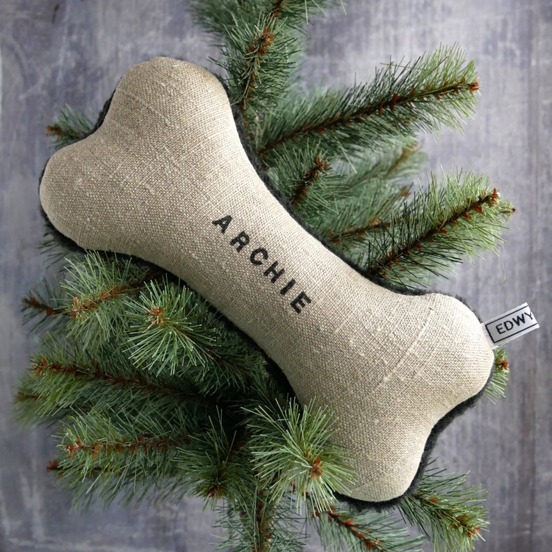 Puede incluir: Un juguete para perro en forma de hueso de lino beige con costuras negras y el nombre "Archie" bordado en &eacute;l. El juguete est&aacute; colgado de una rama de pino verde.