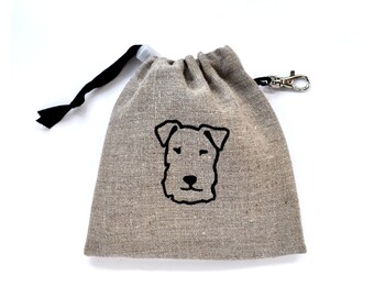 Puede incluir: Una pequeña bolsa de cordón de lino de color marrón claro con una silueta de perro negra y un cordón de ajuste negro. La bolsa tiene un clip de metal plateado.