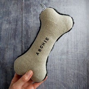 Puede incluir: Un juguete para perro de hueso de lino beige con ribete de piel sint&eacute;tica negra. El juguete est&aacute; personalizado con el nombre "ARCHIE" en letras negras.