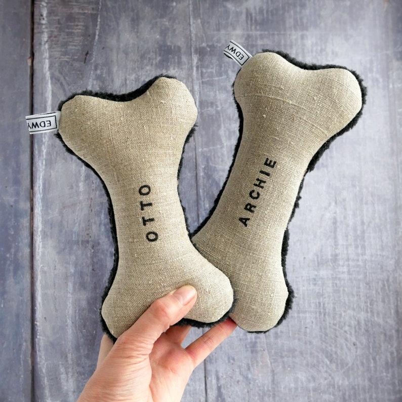 Puede incluir: Dos juguetes para masticar para perros de color beige con forma de hueso y ribete de piel negra. Los huesos est&aacute;n personalizados con los nombres "Otto" y "Archie".