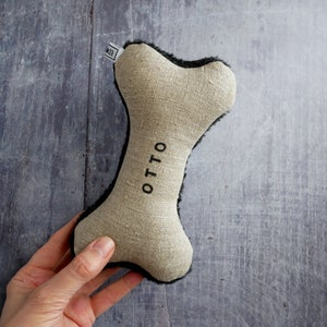 Puede incluir: Un juguete para perro en forma de hueso de lino beige con ribete de piel sint&eacute;tica negra y el nombre "OTTO" bordado en la parte delantera.
