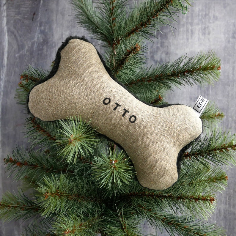 Puede incluir: Un juguete para perro en forma de hueso de lino beige con ribete negro y el nombre "OTTO" bordado. El juguete est&aacute; sobre un &aacute;rbol de Navidad artificial verde.