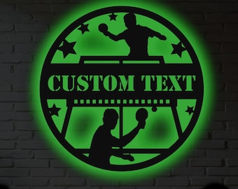 May include: Black metal wall art featuring a ping pong design with a green neon glow. The design includes two players, a table, stars, and the words "CUSTOM TEXT". The art is circular and mounted on a white brick wall.