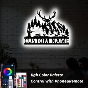May include: Black metal wall art featuring a deer, trees, mountains, and a hunter silhouette. The text "CUSTOM NAME" is displayed below the scene. The image also shows a phone and remote control with the text "RGB Color Palette Control with Phone&Remote".