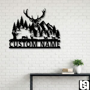 May include: Black metal wall art featuring a deer with large antlers, mountains, and trees. The silhouette includes a hunter and deer. The text "CUSTOM NAME" is at the bottom. The artwork is mounted on a white brick wall.