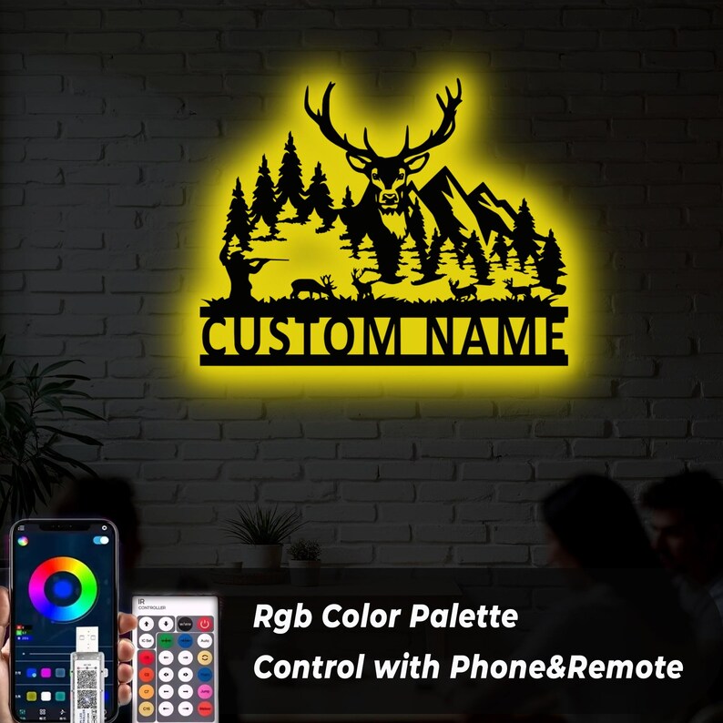 May include: Black metal wall art with a deer silhouette, trees, and mountains, illuminated by a yellow glow. The text "CUSTOM NAME" is displayed below the design. The image also shows a phone and remote control for the RGB color palette.