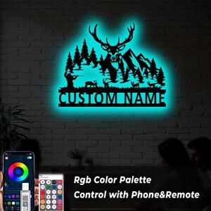 May include: A black metal wall art piece featuring a deer, mountains, and trees, with the text "CUSTOM NAME" illuminated by a teal glow. The image also shows a phone and remote control for color adjustments.