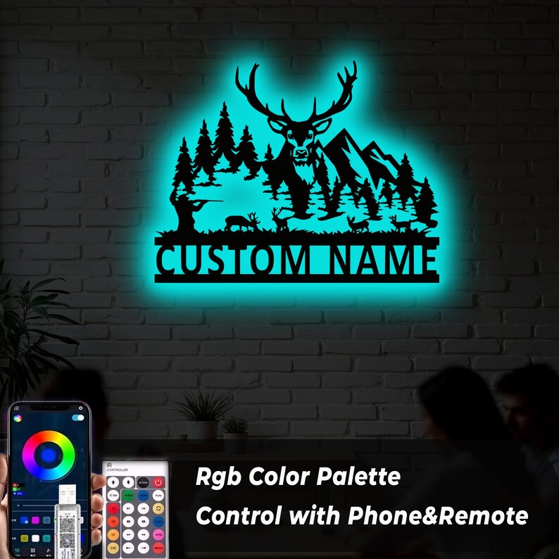 May include: A black metal wall art piece featuring a deer, mountains, and trees, with the text "CUSTOM NAME" illuminated by a teal glow. The image also shows a phone and remote control for color adjustments.