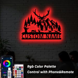 May include: A black metal wall art piece featuring a deer silhouette with antlers, trees, mountains, and the text "CUSTOM NAME" illuminated by a red LED glow. The image also shows a phone and remote control for the RGB color palette.