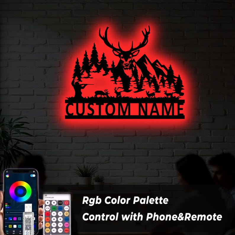 May include: A black metal wall art piece featuring a deer silhouette with antlers, trees, mountains, and the text "CUSTOM NAME" illuminated by a red LED glow. The image also shows a phone and remote control for the RGB color palette.