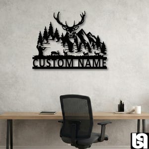 May include: Black metal wall art featuring a deer with large antlers, mountains, and trees. A hunter is depicted in the forest. The text "CUSTOM NAME" is below the scene. The art is displayed on a light gray wall.
