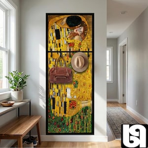 May include: A wall-mounted coat rack featuring a reproduction of Gustav Klimt's "The Kiss" painting. The rack holds a brown handbag and a tan fedora. The artwork is framed in black, with a yellow and gold colour scheme.
