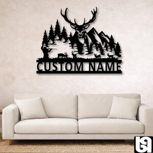 May include: Black metal wall art featuring a deer with large antlers, set against a mountain and forest backdrop. The design includes a hunter and the text "CUSTOM NAME" below the scene. A decorative piece for home decor.