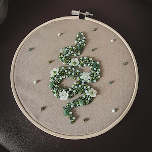 Könnte beinhalten: Handgefertigte Stickerei mit einem Schlangenmuster aus grünen und weißen Perlen, verziert mit weißen Blüten. Das Design ist auf beigem Stoff in einem Holzrahmen angebracht. Streuende Perlen ergänzen das dekorative Kunstwerk.