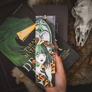 May include: A hand-painted bookmark featuring a gothic illustration of a woman with green and black hair, a snake, and a decorative arch. The bookmark has a gold tassel and is set against a backdrop of books.
