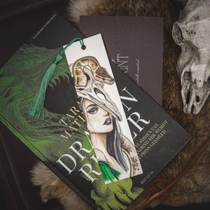 Könnte beinhalten: Aquarell-Lesezeichen mit einer Frau mit Totenkopf-Kopfschmuck und Schlangenmuster. Das Lesezeichen hat eine grüne Quaste und liegt auf einem Buch mit dem Titel "Dragon Rider". Das Bild hat ein dunkles Fantasy-Thema.