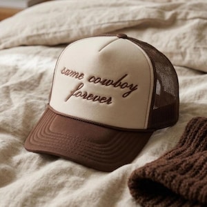 Könnte beinhalten: Braune und beige Trucker-Cap mit dem gestickten Text "same cowboy forever". Die Cap hat eine Mesh-Rückseite und einen gebogenen Schirm. Die Cap liegt auf einer beigen Oberfläche.