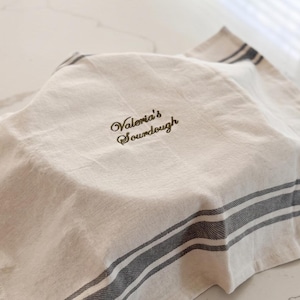 May include: A beige kitchen towel with dark gray stripes along the edges, draped over a round object. The fabric features the embroidered text "Valeria's Sourdough" in a dark brown script font centered on the top surface of the cloth.