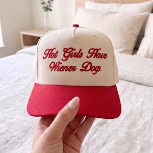 May include: A cream-colored baseball cap with a red brim. The phrase "Hot Girls Have Wiener Dogs" is embroidered in red script across the front of the hat.