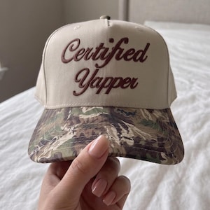 May include: A beige baseball cap featuring the text Certified Yapper embroidered in dark brown script on the front crown. The cap has a structured five-panel design and a curved brim with a green, brown, and black camouflage pattern print.