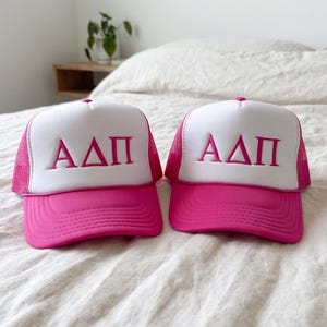May include: Two pink and white trucker hats with the Greek letters ΑΔΠ embroidered on the front. The hats have a pink brim and mesh back, with a white front panel. The hats are displayed on a bed.