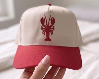 Peut inclure: Casquette de baseball avec une couronne crème et une visière rouge. Le chapeau présente un motif de homard rouge brodé sur le devant. Ce chapeau est de style classique, adapté au port décontracté.