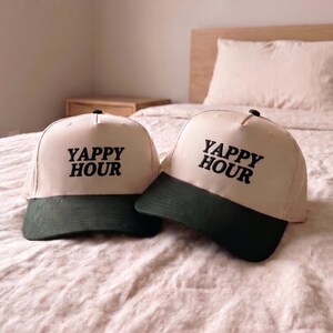 May include: Two baseball caps with a cream-colored crown and a dark green brim. The text "YAPPY HOUR" is printed in black on the front of each cap. The caps are resting on a bed with a pink comforter.