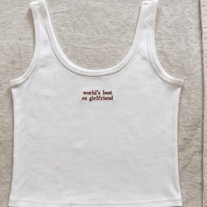 May include: A white ribbed tank top featuring a scoop neckline and thick shoulder straps. The center of the chest displays the text "world's best ex girlfriend" embroidered in a small, dark red, sans-serif font on the fabric surface.