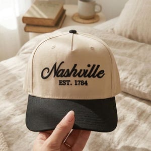 May include: A tan baseball cap with a black brim. The cap features the word "Nashville" embroidered in black script, with "EST. 1784" below it. The cap is held by a hand.