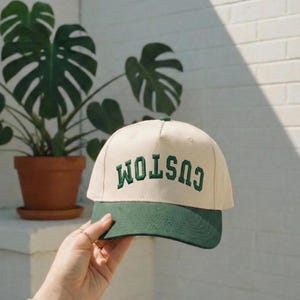 Op de afbeelding: Crème en groene baseballcap met het woord "CUSTOM" in het groen geborduurd. De pet wordt vastgehouden voor een potplant met grote groene bladeren.