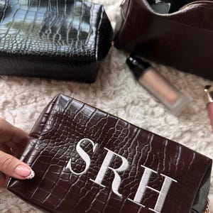 May include: A collection of brown and black faux-leather cosmetic bags with a crocodile skin texture. One bag is embroidered with the initials "SRH" in white. Makeup products are visible in the background.